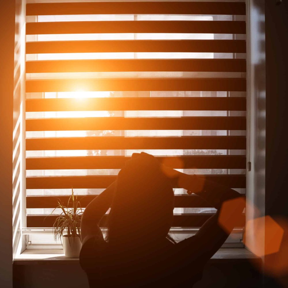 bright sunbeam through gap in blind illuminates silhouette of girl sitting on bed in bedroom and braiding ponytail on her head